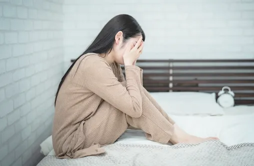 ベッドの上で膝を抱え、顔を手で覆っている女性。ストレスや悩みを抱えている様子が感じられる室内の場面。