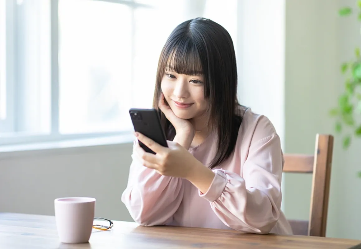 スマートフォンを見ながら微笑む若い女性。明るい室内でリラックスした雰囲気が伝わる。