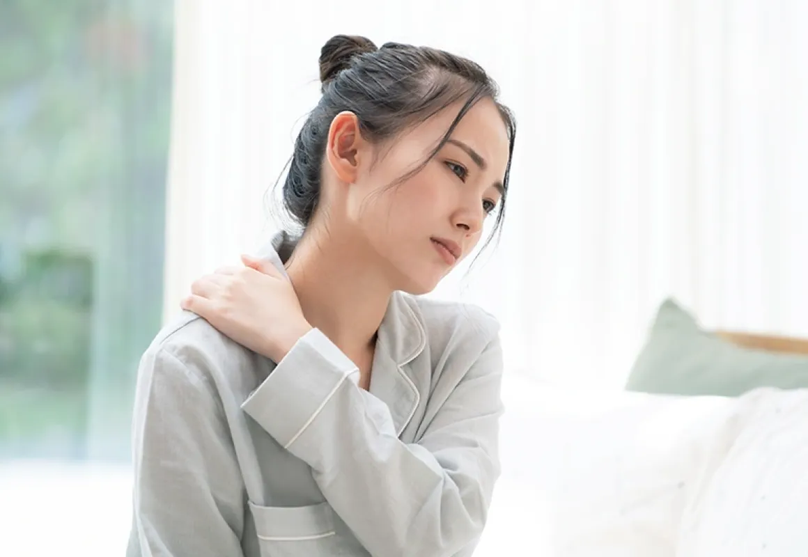 肩を押さえながら疲れた表情を浮かべる女性。室内でリラックスした服装をしている。