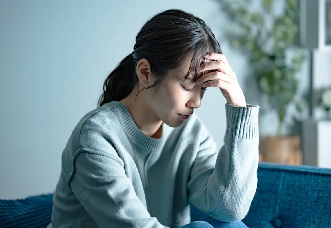 ソファに座り、頭に手を当てて悩んでいる様子の女性。ストレスや疲労を感じているような静かな室内の場面。