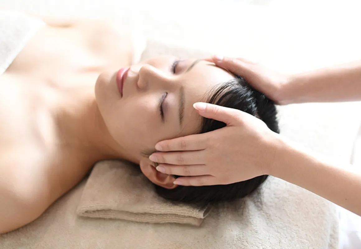 リラクゼーションマッサージを受けている女性。タオルに頭を乗せ、目を閉じてリラックスした表情で、頭部に施術を受けている様子。